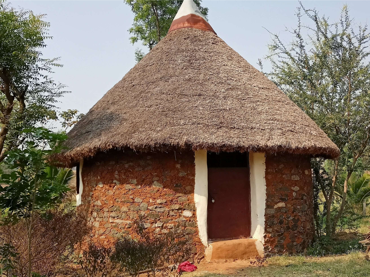 A 16 feet diameter round hut with stone and cob walls and local rough wood and thatch roof, no belt beam, mud flooring, metal door and window-  Total area 200 square feet; total cost Rs 150000/-